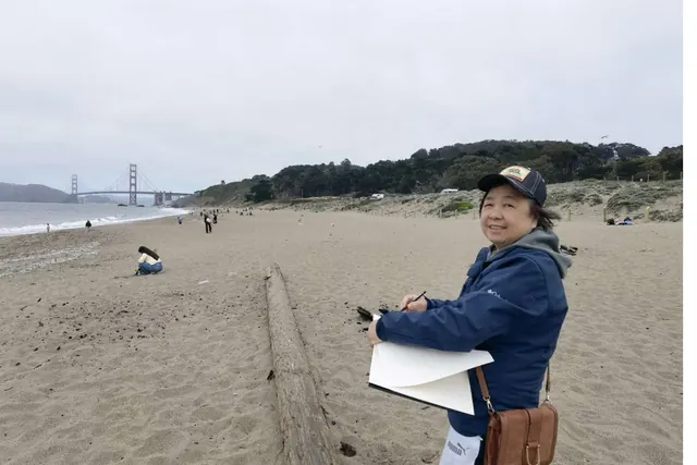 Mary Liu painting at her easel on the beach, with the Golden Gate Bridge behind her